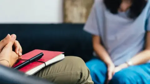Getty Images A person resting their hands on a notebook cross-legged, and a woman pictured in the background out of focus.