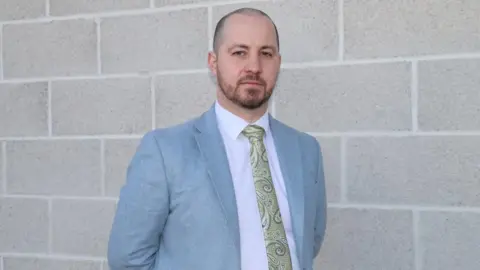 Hertfordshire Police Det Ch Insp Nick Gardner: a man with short dark hair and dark beard with his hands clasped behind his back. He is wearing a pale blue jacket, pale shirt and yellow patterned tie.