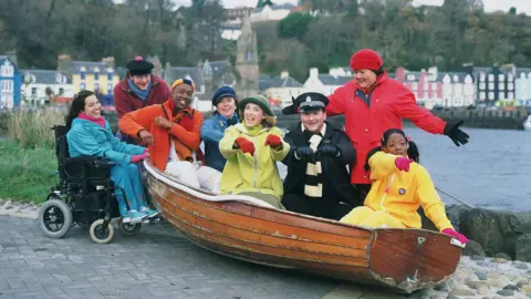 BBC The cast of the original Balamory pose with a wooden rowing boat across the bay from the colourful facades of the houses in Tobermory on Mull