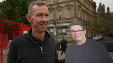 Spencer Stokes, the reporter, stands holding a cut out of Andy Burnham in Huddersfield.