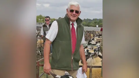 Thame Farmers Auction Mart Richard Roff, chairman at Thame Farmers Mart, in a pen among black and white goats. He is holding a piece of paper and is wearing shades. Another man also wearing shades can be seen behind him.