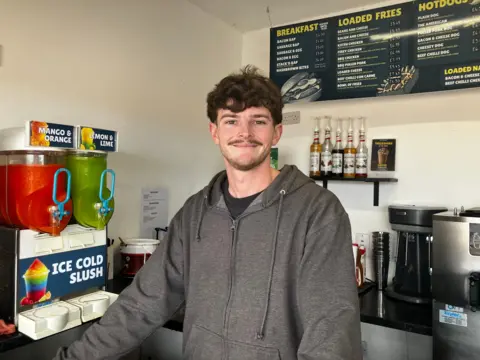 BBC News A man in a grey hooded jumper stands next to a slushy machine and in front of a coffee machine. He is smiling at the camera. 