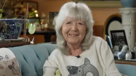 Dame Jilly in her living room on a pale blue sofa smiling to camera