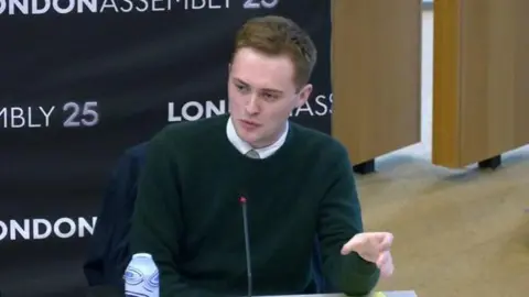 London Assembly A man in a dark green jumper and white shirt sits talking in front of a microphone. 