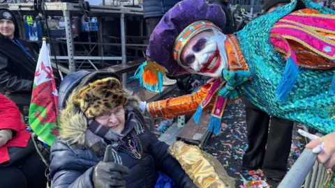 Photograph of Avril Davidge, sat outside in her wheelchair. A Welsh flag can be seen poking out of her wheelchair. She wears a leopard furry hat, a purple scarf and a black puffer coat. She wears small reading glasses and holds her thumb up to the camera. A captain of the Mummers parade leans down to her and smiles. He wears a vibrant blue, purple and orange sequined costume and has a large red joker smile painted on his face. 