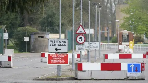 PA Media The image shows the entrance to a former military facility, viewed from a road leading up to a checkpoint area. The entrance is controlled with barriers and multiple warning signs. In the front-left of the image, a rectangular white sign with black text reads: VISITORS VEHICLES with a left-pointing arrow. Below it, a red sign with white text states: DRIVERS TRAVELLING FROM EUROPE TO IDENTIFY THEMSELVES TO SECURITY STAFF