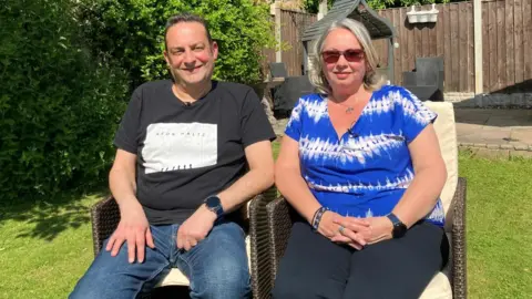 BBC Del and Aly Thornton are sitting side by side on two chairs in their garden. They are both smiling directly at the camera.