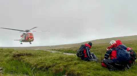 SLMRT responders awaiting the departure of Irish Coast Guard helicopter, Rescue 118. There are three team members in navy and red waterproofs, wearing hats and backpacks. They crouch on the grass. Mountains are visible in the background. The sky is cloudy.

