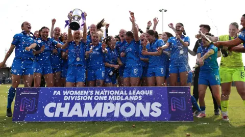 DARREN WILES The team standing in a group on the football pitch and cheering as they are sprayed with champagne. One of them is holding the trophy. In front of them is a sign that says "FA Women's National League Division One Midlands Champions". 