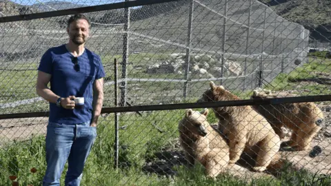 IAR/FPWC Steven Cree wearing jeans and a t-shirt, posing for the camera beside a wire fence. Three light brown bears are sat on the other side of the fence in a large enclosure.