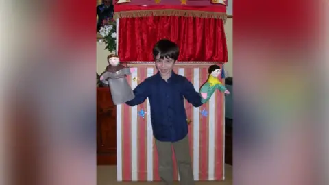 An older photograph of a young person with short brown hair wearing a navy top and green trousers. They are standing in front of a red and white striped puppet theatre, and they have a puppet on each hand