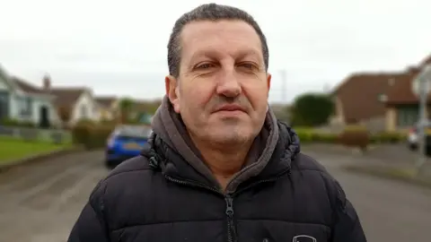 Councillor Wesley Irvine is standing on a street. He has short brown-grey hair and stubble and is wearing a dark padded coat with a maroon hoodie underneath. The street is out of focus behind him.