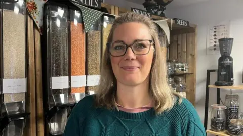 Photo of Caroline, who's looking into the camera and smiling slightly. She's wearing a dark green woollen jumper, and has shoulder-length blodne hair and dark-framed glasses. She's stood in front of a row of containers, which have lentils, seeds, and nuts in - and have openings at the bottom allowing people to refill their own containers. Above the shelving are signs, in white writing on a black background, that read 'Lentils, Snacks, Seeds'