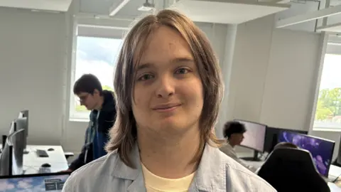 Josh Lovatt/BBC Alfie Johnson is standing in a room where there are several computers. Johnson has shoulder-length brown hair and is wearing a yellow T-shirt and blue shirt.