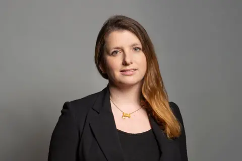 UK Parliament Official portrait of Minister for Employment and MP for Birkenhead, Alison McGovern. She has long brown hair over one shoulder, and is wearing a black suit and gold necklace