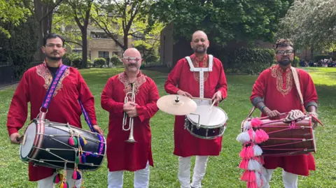 Phil Harrison/BBC Four men in red dress and white trousers carry musical instruments including drums and a trumpet 