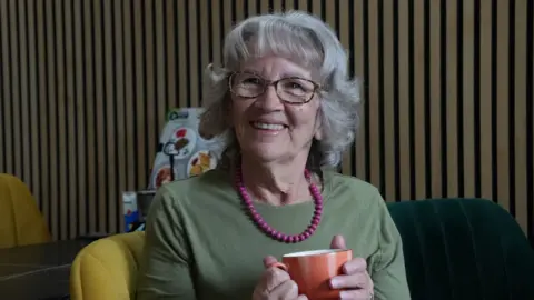Connor Bennett/BBC Margaret Davy siedzi na krześle w restauracji z kubkiem do kawy w rękach. Ma faliste siwe włosy i ma na sobie okulary, zielony top i ma różowy naszyjnik z koralikami