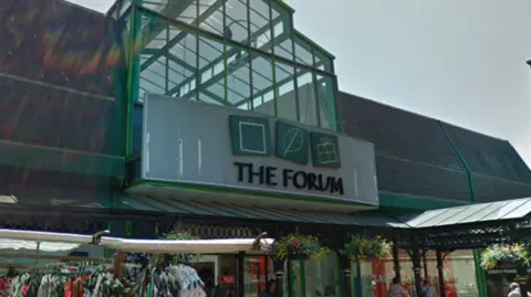 A sign above a shopping centre entrance which reads The Forum in black capital letters. Above the wording are three squares, one depicting a shopping bag, the others with a leaf and a wrapped gift. The sky is grey in the background and hanging baskets are under the sign. 