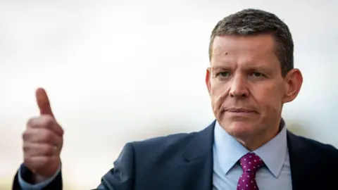 Rhun ap Iorwerth, leader of Plaid Cymru, wearing a navy blue suit with a red shirt and a purple tie, makes the thumbs up gesture as he looks away from the camera 