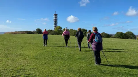 Five people facing away from the camera are walking up a hill at Stoke Park. They all have two poles. A satellite tower, Purdown BT Tower, is seen in the distance, near some trees.