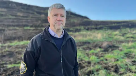 Head and body image of Martin Gray from Historic Environment Scotland. He is standing in front of an area of grassland damaged by the fire on Arthur's Seat. He is wearing at dark zipped fleece with the HES logo on the arm. He has grey hair and a short beard and moustache.