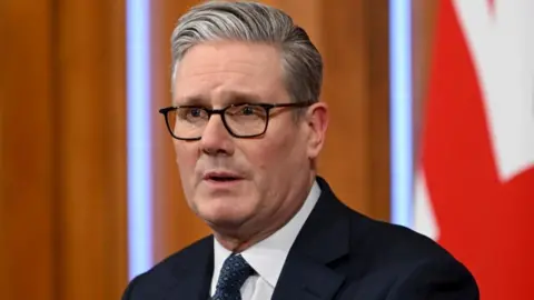 Sir Keir Starmer speaking at a press conference. He is wearing a black suit and navy patterned tie