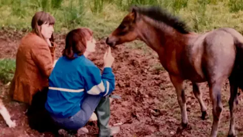 Stella Gratrix Ginny and Stella sitting on the ground next to a small brown foal. Both have a fringe and dark hair, and are turned away from the camera towards the foal. Ginny is wearing a brown jacket while Stella is wearing a bright blue jacket. 