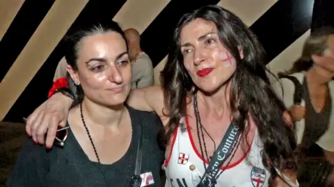 A woman with long dark looks at the camera, wearing red lipstick and with the George cross painted on her cheek.