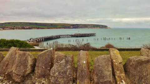 Robin Red Breast A Victorian pier snakes out into the sea. On the horizon are cliffs covered in a patchwork of fields. In the foreground, there are large boulders.