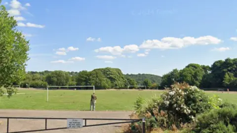 An area of grass with two sets of goalposts and surrounded by trees. In the foreground is a metal gate across an access road.