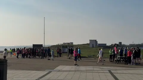 Runners completing the Blyth Parkrun on a sunny day. There are about 100 runners standing recovering and one or two just crossing the line to the far left. The defensive World War Two buildings that make up Blyth Battery can be seen behind the runners.