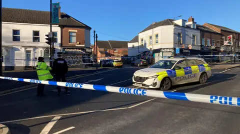 Simon Thake/BBC A street cordoned off with bright yellow police tape blocking access to the road. A police car is parked in the centre and two uniformed officers stand nearby.