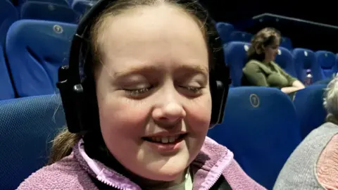 Eryn has long brown hair and is pictured at the cinema in a pink fleece and is wearing an audio descriptive headset. The cinema seats are all blue and a few people are seen in the cinema in the background. 