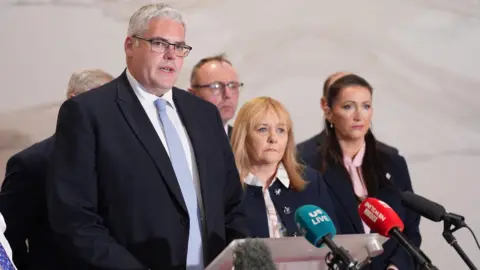 PA Media Gavin Robinson speaking in front of microphones. He has short white hair, wearing a navy blazer, white shirt and light blue tie. Other members of the DUP are standing beside him.