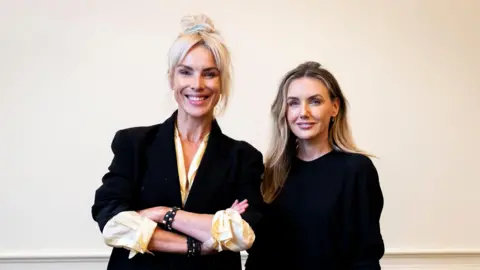 Sali Hughes and Jo Jones standing next to each other, with Hughes's arm linked through Jones's. They are both looking at the camera, wearing dark clothes and smiling.