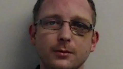 Man with short hair and glasses looking at the camera against a plain light-coloured wall.