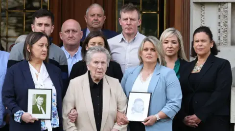 A group of people stand together outside a building with pillars. Some of the people are holding framed photos of a man.