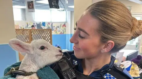 A policewoman holding Larry the lamb and smiling at him at what looks like a police HQ.