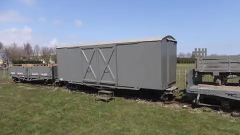 Chris Bates/LCLR A grey 1917 emergency ambulance van with double doors sits on a light-gauge railway track between smaller supply wagons. A small step offers access to a grassy field