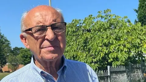 Terry Yems is wearing glasses and a light blue shirt and is standing beside a metal fence and leafy trees on a bright sunny day.