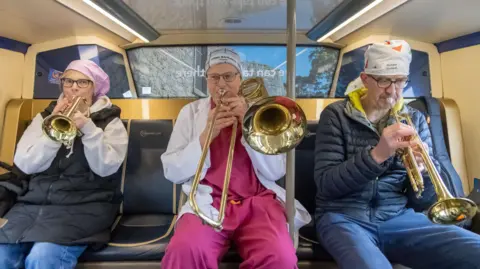 Supplied Three people are sitting on a bus playing brass instruments, including trumpets and a trombone. The bus interior, windows, and overhead lighting are visible in the background. They are wearing reusable hats. 