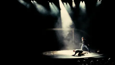 Scene from the play Punch, on the West End, spotlight is on two men, one that is on all fours, the other standing over him. 