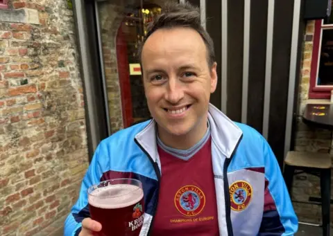 Stewart Garratt Stewart Garratt, wearing an old fashioned claret Villa shirt and a claret and blue tracksuit top while holding a pint of beer.  He is seated outside by a brick wall.