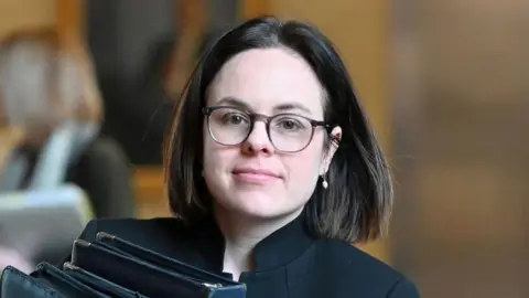 Getty Images Kate Forbes has short dark brown hair and wears glasses and a black top while carrying black folders
