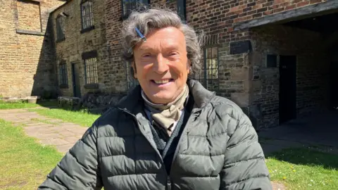 BBC/Victoria Scheer Harrison sits in bright daylight in front of a group of historic stone and brick buildings. He is wearing a padded, quilted jacket in a muted green tone and a clip in his hair.
The buildings behind him have weathered brickwork, stone walls, small multi‑pane windows, and dark-painted window frames, giving the setting an industrial or early historic appearance. A paved path runs through a grassy courtyard area, with patches of sunlight and shadows.