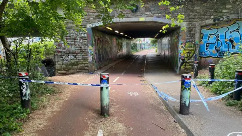 BBC Blue and white police tape runs between bollards blocking a railway bridge. A red cycle path runs underneath the bridge, and is blocked off by police tape.
