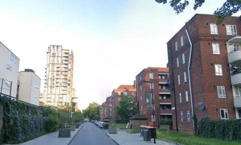 A small residential road is pictured going through Woodberry Down Estate which has several five-floored flats running down the righthand side of the picture and a tower block in the distance on the left