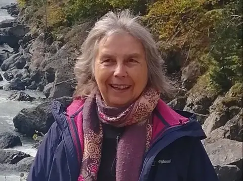 Anne Page Anne Page has grey hair and a smile. She is wearing a purple patterned scarf and a purple coat. She is standing by a rocky sea shore.