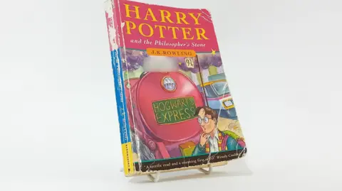 Plymouth Auction Rooms A paperback book which is worn at the edges and has a dog eared corner bears the words Harry Potter and the Philosopher's Stone. It also has a sticker saying 'I belong to' on the front. 