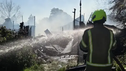 firefighter in a fluorescent helmet aims a hose into a smoked building where corrugated iron sheets and fire-damaged wreckage can be seen